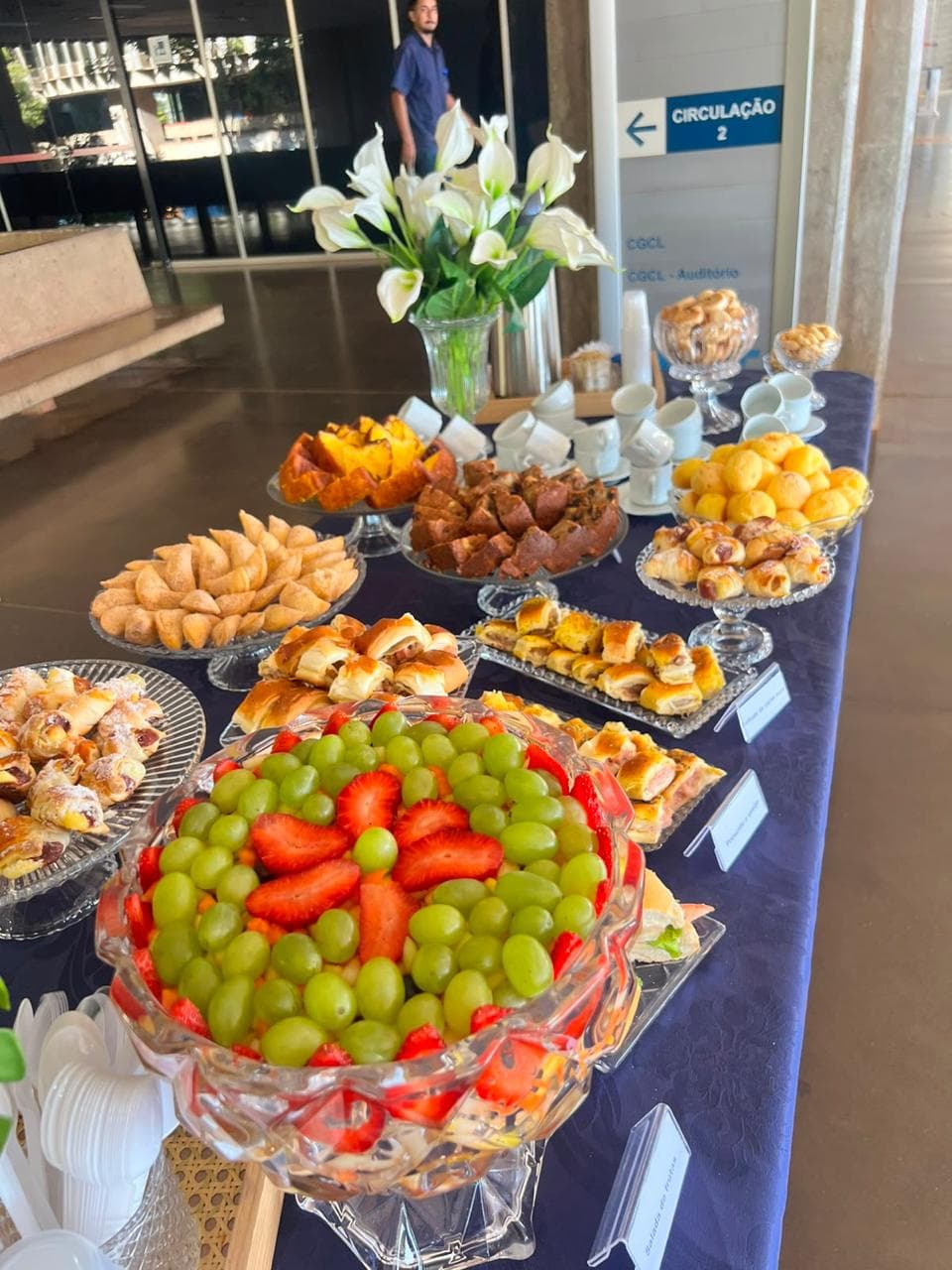 Mesa com toalha azul, salada de frutas em cristal, folhados e xicaras de cafe