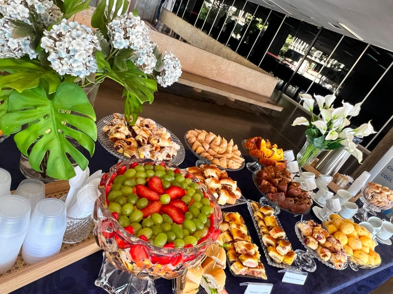 Mesa de coffee break com arranjos de flores brancas e travessas de cristal
