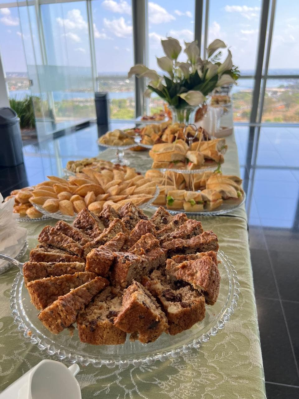 Bolo em fatias, pasteis e sanduiches em mesa diante de janela panoramica